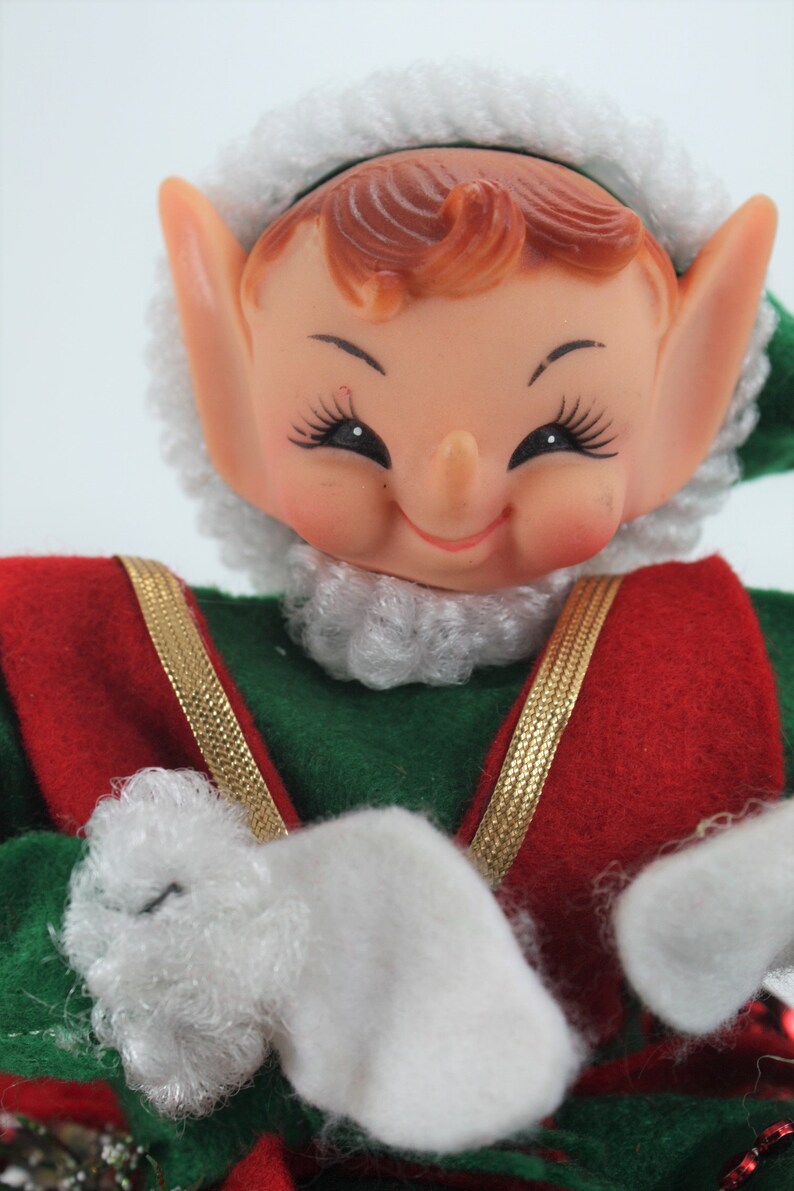 May include: A smiling elf doll with a white fur hat and beard, wearing a green and red outfit with gold trim. The elf is holding its hands in front of it, with white fur mittens.