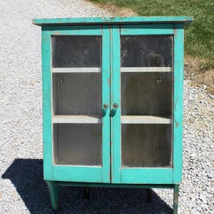 May include: A teal blue cabinet with glass doors and shelves. The cabinet has a distressed finish and simple metal knobs.