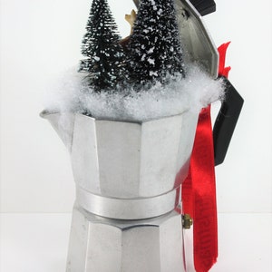 May include: A silver metal coffee pot with a red ribbon and a snowy winter scene inside. The scene includes two small artificial pine trees and a small deer figurine.