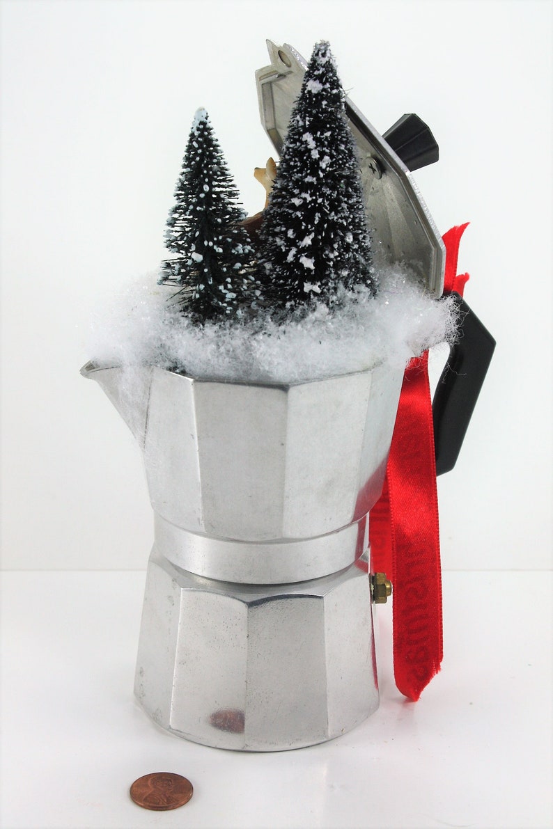 May include: A silver metal coffee pot with a red ribbon and a snowy winter scene inside. The scene includes two small artificial pine trees and a small deer figurine.