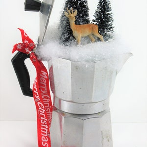 May include: A silver coffee pot with a red ribbon that says "Merry Christmas" tied around the handle. The pot is filled with fake snow and small artificial pine trees and a deer figurine.
