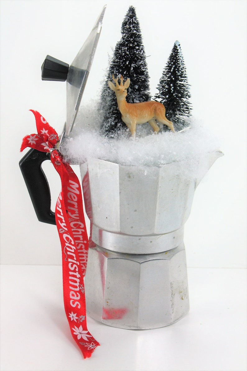 May include: A silver coffee pot with a red ribbon that says "Merry Christmas" tied around the handle. The pot is filled with fake snow and small artificial pine trees and a deer figurine.