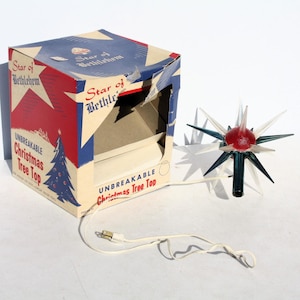 May include: A Star of Bethlehem Christmas tree topper with a red center and radiating teal and clear plastic spikes. The item includes a white electrical cord and plug, and is presented next to its original cardboard box with blue and red graphics.