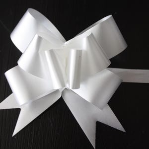 May include: A bright white decorative pull bow made of smooth, satin-like ribbon. The bow features multiple layered loops arranged in a circular pattern with two pointed, V-shaped tails extending downward and a single flat ribbon tail extending right.