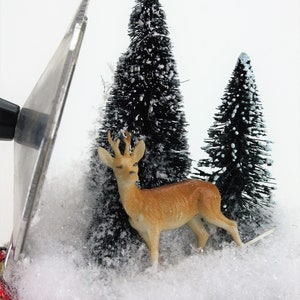 May include: A miniature winter scene with a brown deer figurine standing in front of two small, snow-covered pine trees. The scene is set in a white, snow-covered base with a red ribbon.