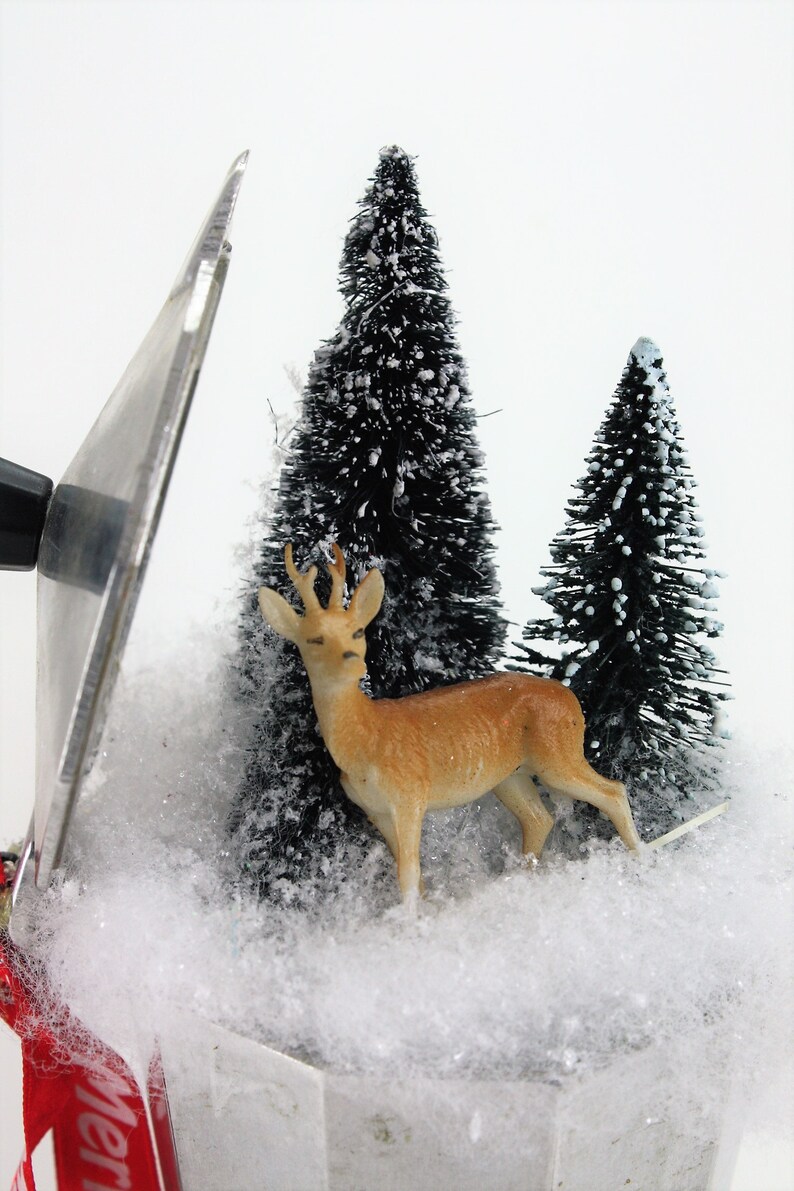 May include: A miniature winter scene with a brown deer figurine standing in front of two small, snow-covered pine trees. The scene is set in a white, snow-covered base with a red ribbon.