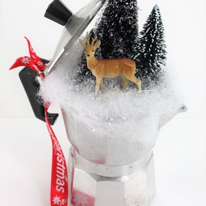 May include: A silver coffee pot with a glass lid decorated with a miniature winter scene. The scene includes two small artificial pine trees, a deer figurine, and white cotton-like snow. A red ribbon with the word "Christmas" is tied to the handle.