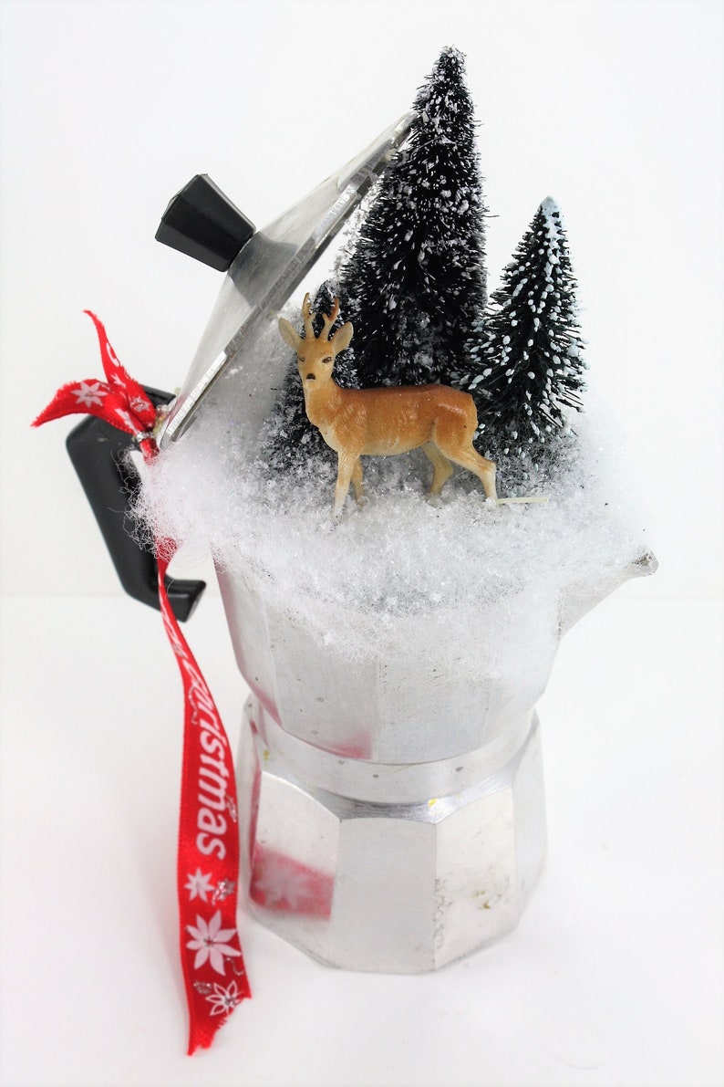 May include: A silver coffee pot with a glass lid decorated with a miniature winter scene. The scene includes two small artificial pine trees, a deer figurine, and white cotton-like snow. A red ribbon with the word "Christmas" is tied to the handle.