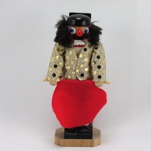 May include: A wooden nutcracker figurine of a matador wearing a gold sequined jacket and holding a red cape. The nutcracker has a black beard, a black hat, and a painted face with a red nose.