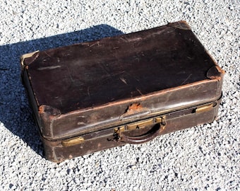 May include: A vintage brown leather suitcase with brass hardware, including corner protectors and a handle. The suitcase shows signs of wear and tear, with scratches and worn edges, suggesting age and use. The suitcase is rectangular and sits on a gravel surface.