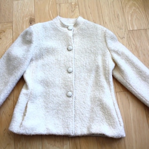 May include: Off-white, textured jacket with a stand-up collar and three buttons down the front. The jacket has long sleeves and two small pockets. The fabric appears to be a soft, woven material.
