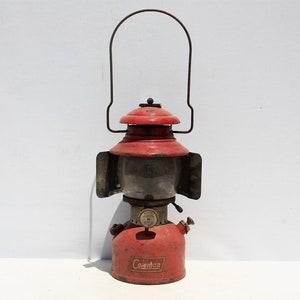 May include: Vintage red Coleman lantern with a clear glass globe and a black handle. The lantern has a red top and base with the Coleman logo. A classic camping or emergency light.