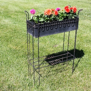 May include: Black metal plant stand with two shelves. The top shelf holds a black planter with pink and orange flowers.