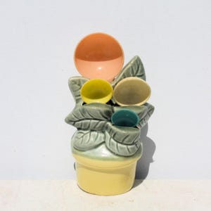 May include: A ceramic measuring spoon set shaped like a potted plant with sage green leaves and a yellow base. The set includes four round measuring spoons in peach, yellow, cream, and teal, which are nested within the green leaf structure.