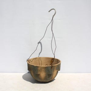 May include: A hanging planter with a natural, earthy aesthetic. The bowl-shaped planter is made of a light brown material with green accents, and is suspended by a wire and a metal hook. The design suggests a rustic or outdoor theme.