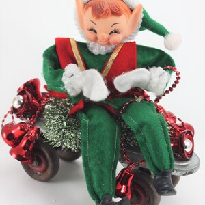 May include: A green and red elf doll riding a toy skateboard decorated with red jingle bells and green tinsel.