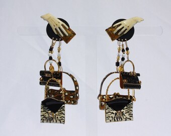 May include: A pair of dangle earrings with a gold chain and three miniature purses. The top purse is black with gold accents, the middle purse is brown and white leopard print, and the bottom purse is black with a white and black zebra print. The top of each earring features a black disc with a white hand holding a gold clutch.