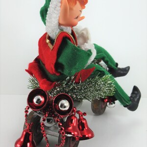 May include: A vintage Christmas elf doll riding a toy wagon with red bells and a green and red outfit. The elf has a white fur hat and closed eyes.