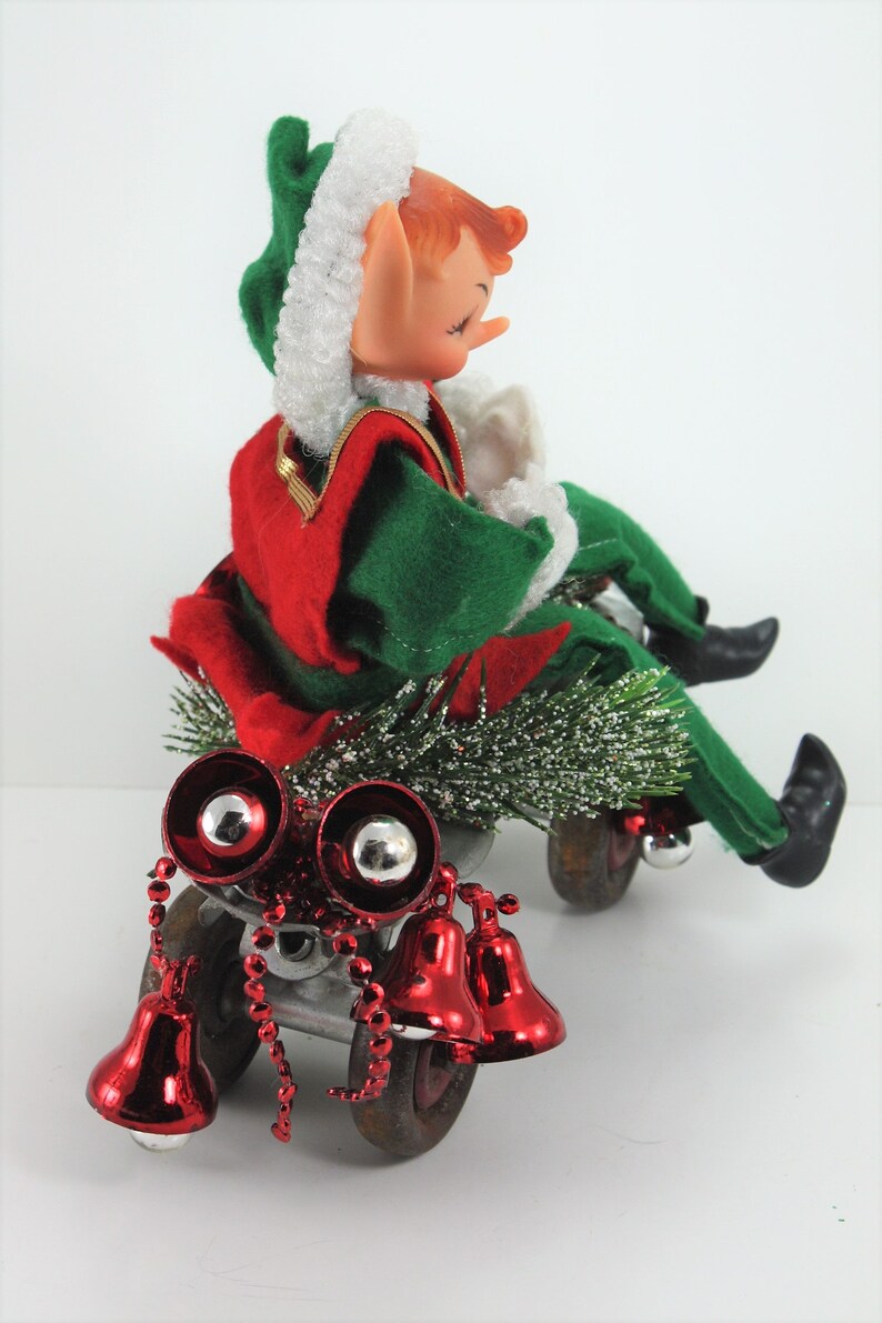 May include: A vintage Christmas elf doll riding a toy wagon with red bells and a green and red outfit. The elf has a white fur hat and closed eyes.