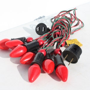 May include: A string of red Christmas lights with black bases and red and green wiring. The lights are arranged in a cluster, with a black plug visible. A yellow tag is attached to the wiring.