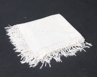 May include: A white crocheted throw blanket with a fringe border. The blanket is folded and lying on a black surface.