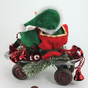 May include: A green and red elf doll riding a toy wagon decorated with red bells and green pine branches.