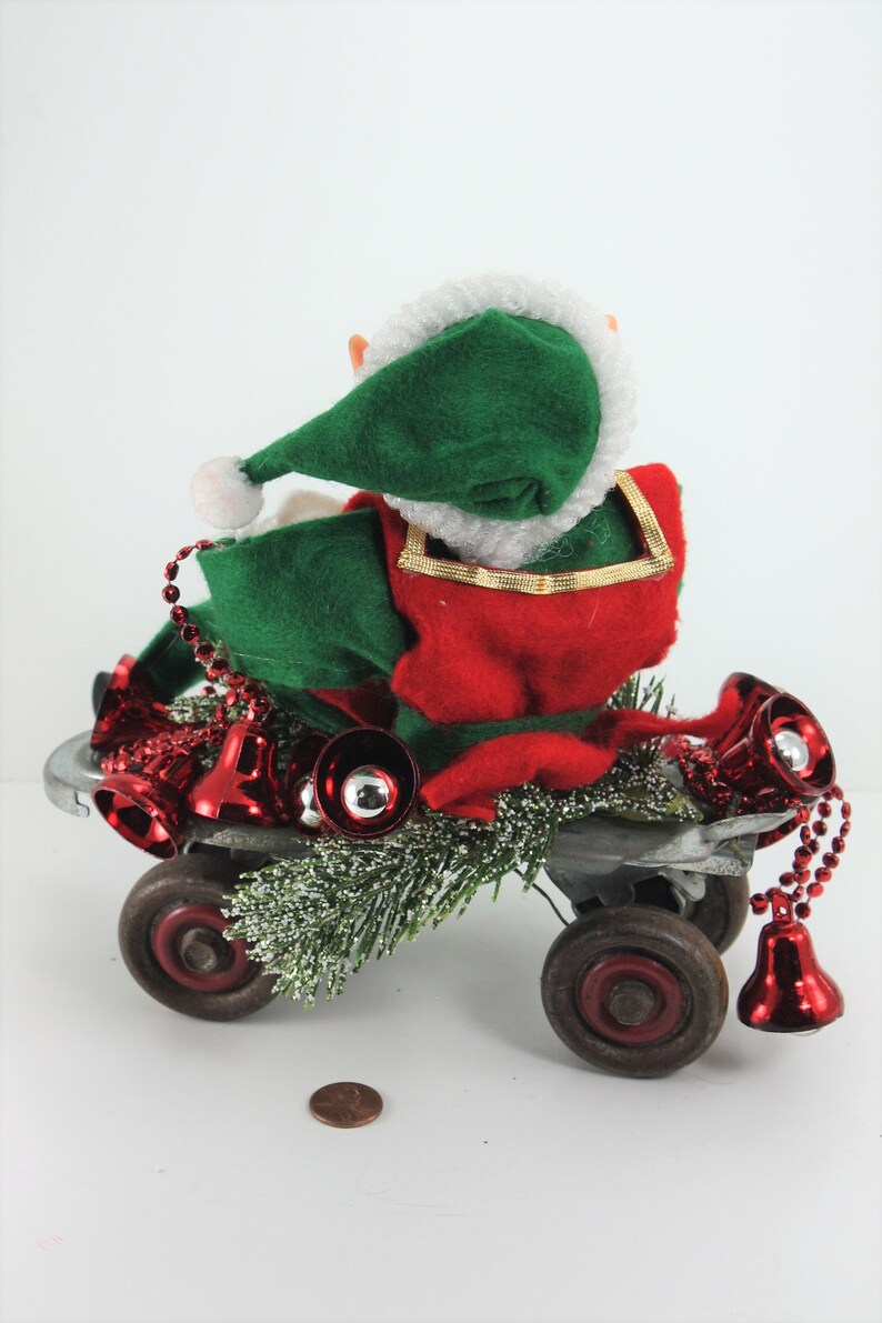May include: A green and red elf doll riding a toy wagon decorated with red bells and green pine branches.
