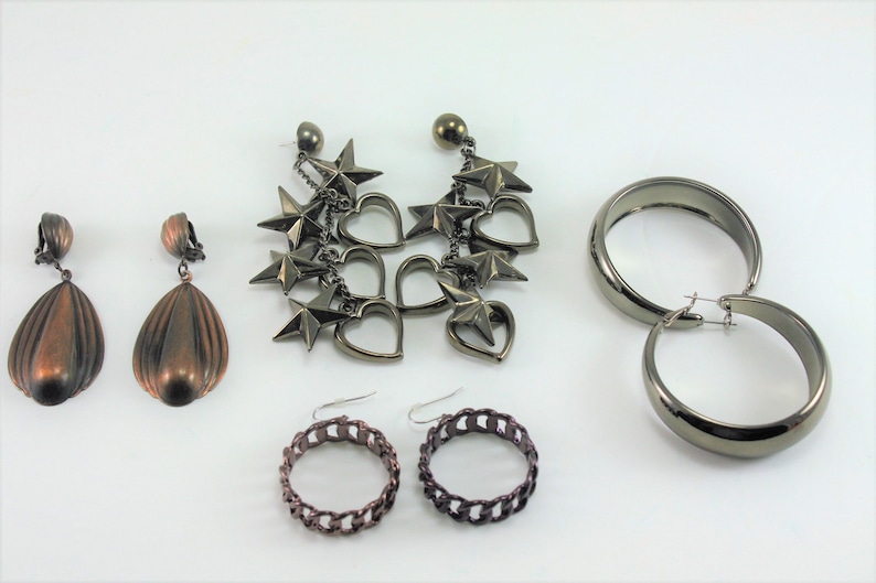 May include: Five pairs of earrings in various styles. Two pairs are copper-colored and have a teardrop shape. One pair is silver-toned and features a dangling chain with stars and hearts. The other two pairs are silver-toned hoops, one pair is thick and the other is a chain link style.