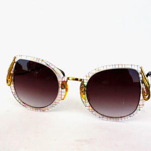 May include: Sunglasses with a white frame featuring a grid pattern in multiple colors. The lenses are gradient brown. Gold-colored decorative accents are on the temples.