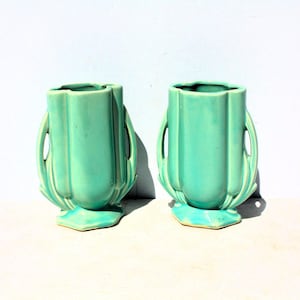 May include: A pair of matching turquoise ceramic vases featuring a vertical ribbed design, a wide open top, and two integrated side handles that curve downward to meet the base. The vases have a smooth, glossy finish and a hexagonal foot.