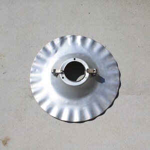 May include: A silver metal lampshade with a fluted edge and two metal clips. The lampshade has a circular opening in the center.