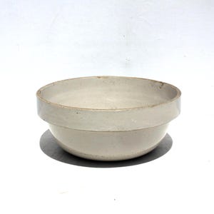 May include: A round, off-white ceramic bowl with a smooth, glazed interior and a distinct, wide horizontal rim around the upper edge. The exterior surface features a subtle speckled texture throughout, with a slightly darker, earthy tone along the rim.