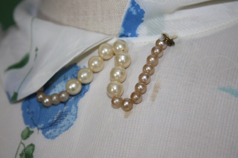 May include: A white blouse with a blue floral pattern and a white collar. A pearl necklace is attached to the collar with a small metal clasp.