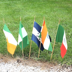 May include: Five small flags on wooden poles are planted in the grass. The flags represent different countries: Ireland, Nigeria, Honduras, India, and Mexico. The flags are colorful and waving slightly in the breeze.