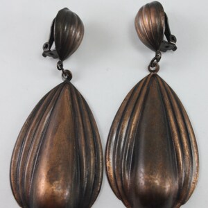 May include: A pair of large, teardrop-shaped, bronze-colored clip-on earrings. The earrings have a ridged, textured surface.