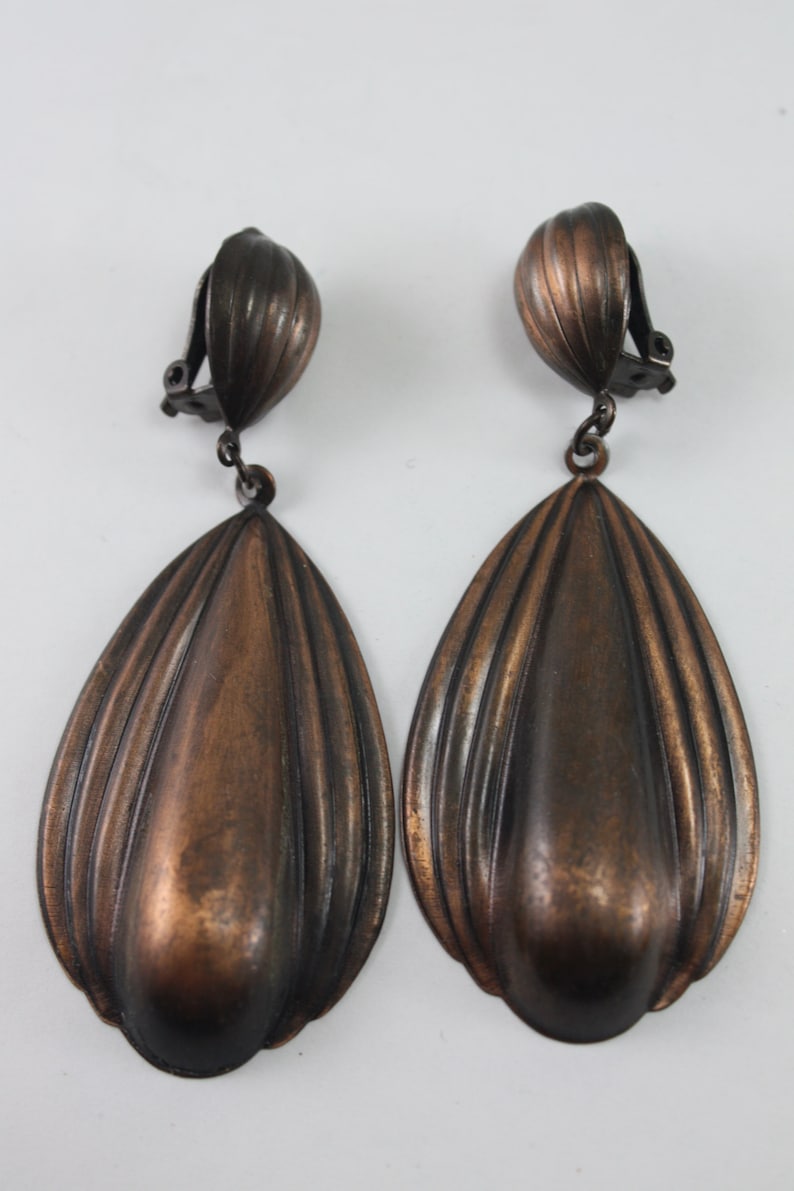 May include: A pair of large, teardrop-shaped, bronze-colored clip-on earrings. The earrings have a ridged, textured surface.