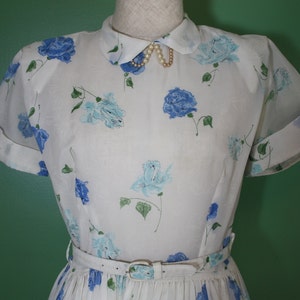 May include: A white dress with a blue floral print and a white collar with a pearl necklace. The dress has short sleeves and a fitted bodice. The skirt is pleated and falls to the knees.