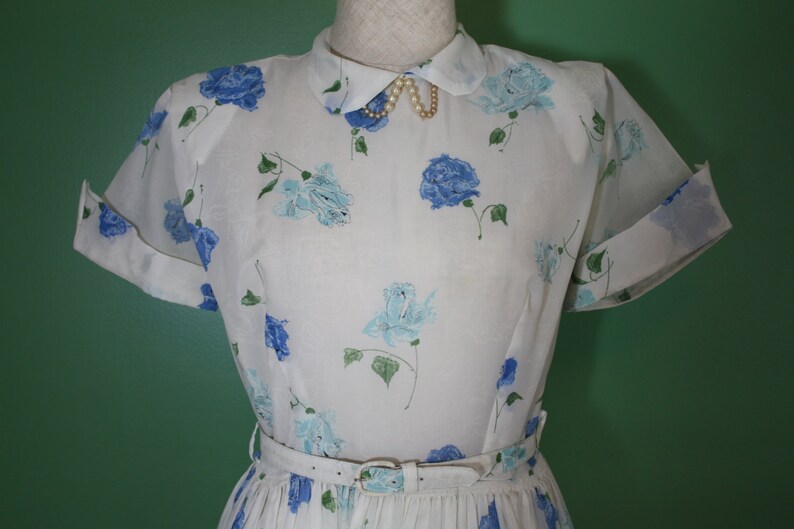 May include: A white dress with a blue floral print and a white collar with a pearl necklace. The dress has short sleeves and a fitted bodice. The skirt is pleated and falls to the knees.