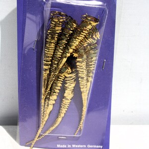 May include: A set of four gold-colored metallic icicle ornaments arranged vertically on a purple cardboard backing. Each icicle features a spiraled, tapered design with a reflective finish. The bottom of the packaging reads Made in Western Germany.