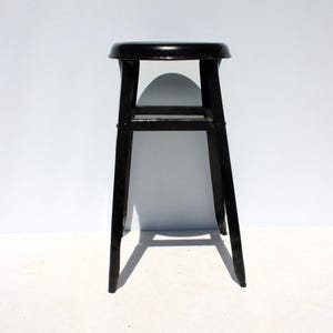 May include: A black metal stool featuring a round seat and four straight legs connected by a square support frame. The stool has a glossy finish and a simple, functional design with a sturdy construction, standing upright on a flat surface.