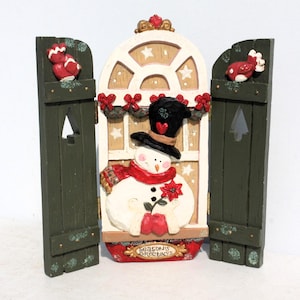 May include: A wooden Christmas decoration featuring a snowman with a red scarf and a red flower in a window with green shutters. The window has a white frame and a brown background with white stars. The text "Season's Greetings" is on the bottom of the window.