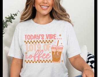 Op de afbeelding: Lichtgrijs t-shirt met de tekst "TODAY'S VIBE... LOTS OF coffee" in roze en oranje. De afbeelding bevat een koffiekopje met een rietje. Het t-shirt wordt door een persoon gedragen.