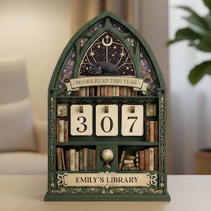 Könnte beinhalten: Ein grüner, holzgeschnitzter Ewiger Kalender in Bücherregalform mit der Aufschrift "BOOKS READ THIS YEAR" und "EMILY'S LIBRARY". Der Kalender zeigt die Zahl "307" und hat eine schlafende schwarze Katze, einen Globus und Miniaturbücher.