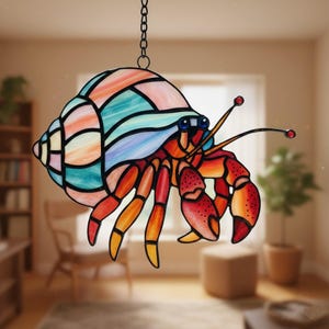 Peut inclure: Un capteur de soleil en vitrail représentant un bernard-l'ermite. Le crabe a une coquille colorée avec des nuances de bleu, de rose et d'orange. Son corps est rouge et orange avec des yeux bleus et de longues antennes. Il est suspendu à une chaîne noire.