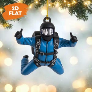 Könnte beinhalten: Blauer Fallschirmspringer-Ornament mit dem Namen "DANIEL" auf dem Helm. Das Ornament zeigt den Fallschirmspringer im Freifall, mit erhobenem Daumen. Das Ornament hängt an einer goldenen Schnur, perfekt für die Weihnachtsdekoration.