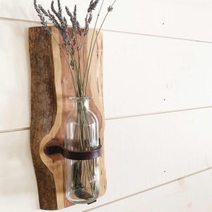 May include: A rustic wall-mounted vase featuring a clear glass bottle held by a dark brown metal band, mounted on a natural wood slab. The vase holds dried lavender stems, creating a natural and decorative display. The wood slab has a live edge.