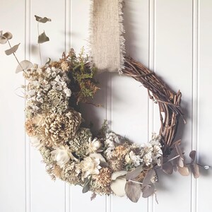 May include: A circular wreath constructed from brown twisted twigs, adorned with a dense arrangement of dried cream, beige, and white flowers, green moss, and eucalyptus sprigs. A wide, frayed-edge tan burlap ribbon hangs from the top.
