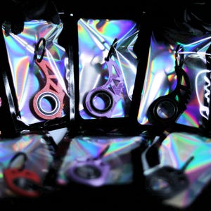 May include: Keychains in red, purple, and black, each with a circular design and a key ring, are displayed in iridescent pouches. The keychains have a unique, geometric shape. A pink skull is visible in the background.