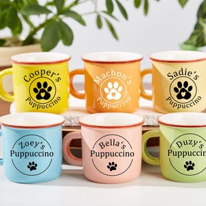 Pode incluir: Seis canecas de cerâmica coloridas com bordas e alças brancas. Cada caneca tem uma cor diferente: amarelo, laranja, amarelo claro, azul, rosa e verde. Cada caneca tem um nome, a palavra "Puppuccino" e um design de pegada.