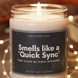 Könnte beinhalten: Eine brennende Kerze in einem Glas mit einem schwarzen Etikett mit der Aufschrift "Smells like a 'Quick Sync' (That ruined my entire afternoon)." Die Kerze steht auf einem runden Holzuntersetzer, umgeben von Lichterketten.